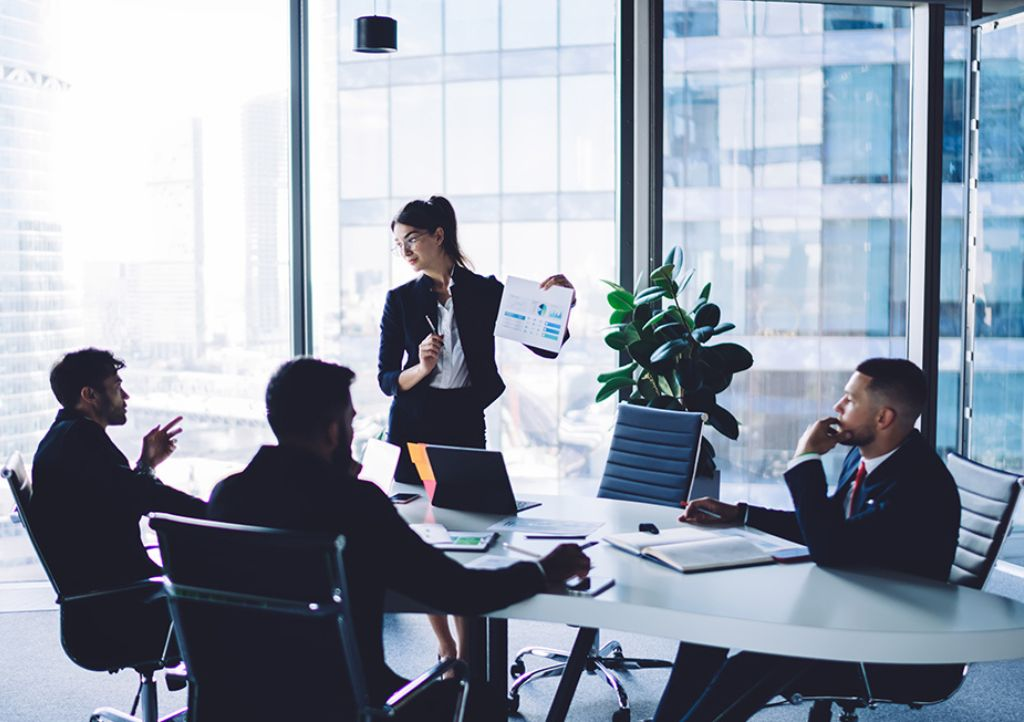 Business meeting in a glass-walled office as a woman presents charts to colleagues at a table.